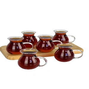 May include: A set of six clear glass tea cups filled with dark tea, each with a silver handle. The cups sit on a rectangular wooden serving board. The tea cups have a rounded base and a slightly flared rim.