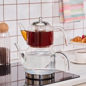 May include: A clear glass teapot with a stainless steel base and lid. The teapot is filled with water and tea, and has a curved handle and spout. The teapot is sitting on a black surface.