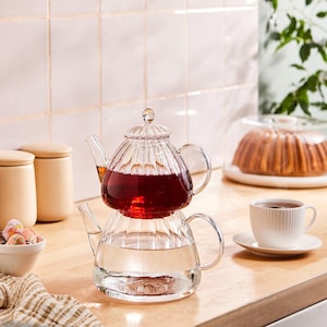 May include: A clear glass teapot with a two-tiered design, filled with red liquid in the top section and clear liquid in the bottom. A white teacup and saucer are on the right. The teapot sits on a wooden surface.