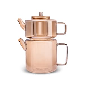 May include: A two-piece, amber-coloured glass teapot set. The bottom pot has a cylindrical shape with a handle, and the top pot, with a lid, sits atop it. The set is designed for brewing tea and has a modern aesthetic.