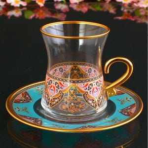 May include: A clear glass teacup with a gold handle and rim, resting on a matching saucer. The cup and saucer feature a teal and gold pattern with red and navy accents. The design is ornate and detailed.