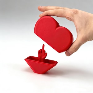 Könnte beinhalten: Ein roter herzförmiger Behälter mit Deckel, der eine rote Hand zeigt, die den Mittelfinger zeigt. Der Behälter besteht aus zwei Teilen, wobei die obere Hälfte angehoben wird. Der Hintergrund ist weiß.
