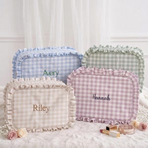 Puede incluir: Cuatro neceseres personalizados en colores pastel: azul claro, verde, beige y lavanda. Cada neceser presenta un estampado de cuadros vichy, un borde con volantes y un nombre bordado en la parte delantera. Los neceseres se exhiben sobre una superficie blanca y suave.
