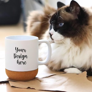 以下が含まれることがあります： コルクベースの白いセラミックマグ。筆記体のフォントで「Your Design here」というテキストが書かれています。茶色、黒、白の毛並みと青い目を持つふわふわの猫が背景にいます。