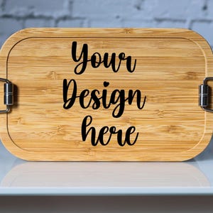 May include: A rectangular bamboo lunchbox with a natural wood grain finish. The lid features the text "Your Design here" in a black, cursive font. The box has metal clasps on each side for closure.