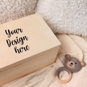 Puede incluir: Una caja de madera de color claro con el texto "Your Design here" en escritura negra en la tapa. Un sonajero gris con forma de oso con un anillo de madera está al lado de la caja. La caja está sobre una manta color crema.