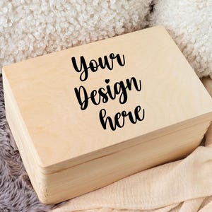 Könnte beinhalten: Eine hellfarbene Holzkiste mit Scharnierdeckel. Auf der Oberseite der Kiste steht in schwarzer Schrift "Your Design here". Die Kiste liegt auf einem beigen Stoff und einer grauen, flauschigen Oberfläche.