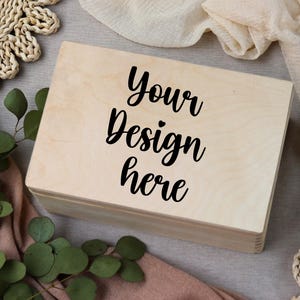 Puede incluir: Una caja de madera de color claro con el texto "Your Design here" en escritura negra. La caja es rectangular y tiene un diseño simple y limpio. La caja está rodeada de vegetación y elementos decorativos.