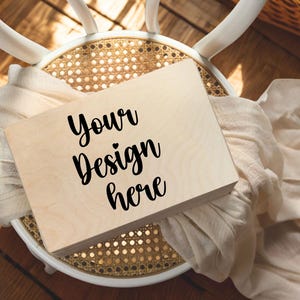 Puede incluir: Una caja de madera de color claro con el texto "Your Design here" en escritura negra. La caja descansa sobre una silla tejida con una tela color crema drapeada sobre ella. La escena está iluminada por la luz natural.