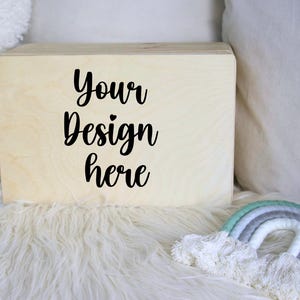 Könnte beinhalten: Eine hellfarbene Holzkiste mit dem Text "Your Design here" in schwarzer Schrift. Die Kiste steht auf einer weißen, flauschigen Oberfläche, mit einer kleinen Regenbogen-Dekoration in der unteren rechten Ecke.