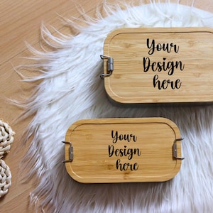May include: Two rectangular bamboo lunch boxes with metal clasps are displayed on a white fluffy surface. The text "Your Design here" is printed in black on the top of each box. The boxes are different sizes.
