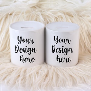May include: Two white ceramic coin banks with a slot on top. Each bank has the text "Your Design here" in black script. The banks are on a fluffy, cream-colored surface.