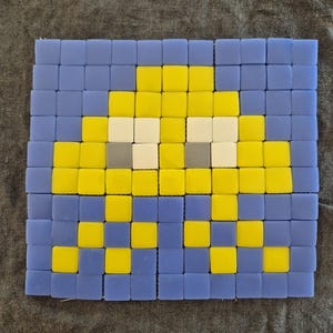 Könnte beinhalten: Ein quadratisches Mosaik mit einem pixeligen Alien-Design. Das Design verwendet blaue, gelbe, weiße und graue quadratische Fliesen. Das Mosaik ist ein Retro-Design, das an klassische Arcade-Spiele erinnert.