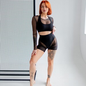 Puede incluir: Conjunto deportivo negro de dos piezas. La parte superior es un sujetador deportivo de estilo halter con mangas de malla transparente. Los shorts de cintura alta tienen paneles de malla transparente. La modelo tiene el pelo rojo y tatuajes.