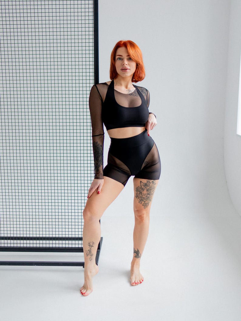 Puede incluir: Conjunto deportivo negro de dos piezas. La parte superior es un sujetador deportivo de estilo halter con mangas de malla transparente. Los shorts de cintura alta tienen paneles de malla transparente. La modelo tiene el pelo rojo y tatuajes.
