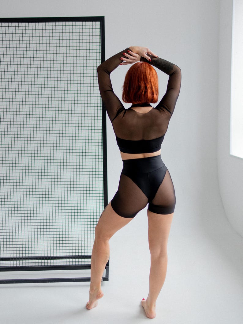 Puede incluir: Conjunto de ropa deportiva negra con una parte superior de malla de manga larga y pantalones cortos de cintura alta con paneles de malla. La modelo tiene el pelo rojo y est&aacute; de pie frente a una rejilla negra.
