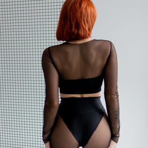 Puede incluir: Conjunto negro de dos piezas con una parte superior transparente de manga larga y pantalones cortos de cintura alta. La parte superior tiene un escote cuadrado. Los pantalones cortos son opacos en la parte delantera y transparentes en los laterales. La modelo tiene el pelo rojo.