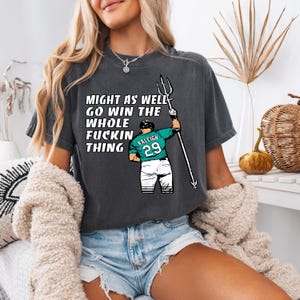 Peut inclure: T-shirt gris foncé avec le texte "MIGHT AS WELL GO WIN THE WHOLE FUCKIN THING". Le t-shirt présente une illustration de joueur de baseball tenant un trident, avec le numéro 29 et le mot "RALEIGH" sur le maillot.