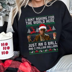 May include: Black sweatshirt with a Christmas-themed design. The design features a man wearing sunglasses and a Santa hat, with text that reads "I AINT ASKING FOR THE WORLD HERE JUST AN 8 BALL AND 2 MILLION DOLLARS."