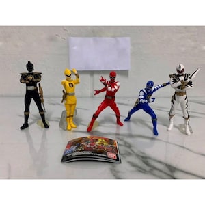 以下が含まれることがあります： 黒、黄色、赤、青、白など、さまざまな色の5体のフィギュアのコレクション。各フィギュアは、詳細なコスチュームとアクセサリーでダイナミックにポーズをとっています。下部には、小さなカラフルなカードがあります。