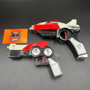 May include: Two toy ray guns, one white, red, and black, the other red, black, and silver. The larger gun has "SPD" printed on the side. A Mahu Suntoy logo is visible on an orange card.