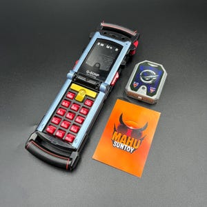 Puede incluir: Un teléfono plegable azul y negro con botones rojos y un botón central amarillo. También hay un dispositivo plateado con un emblema azul y una pequeña tarjeta naranja con el texto "MAHU SUNTOY".