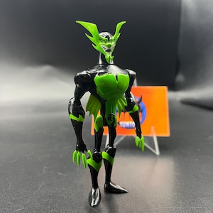 May include: A black and green action figure of a fictional character with a pointed head, clawed hands, and a chest emblem. The figure has green accents and is posed standing upright.