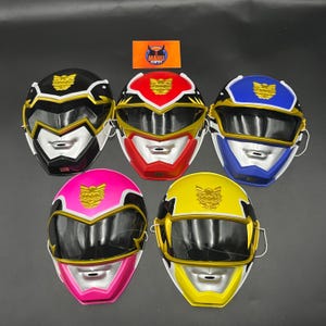 May include: Five Power Ranger masks in black, red, blue, pink, and yellow. Each mask features a gold emblem and visor, with a silver mouth and chin. These plastic masks are designed for cosplay or costume purposes, and are likely for children.