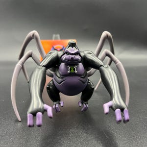 May include: A black and purple action figure of a gorilla-like creature with spider legs. The figure has a green emblem on its chest and purple hands and feet. The figure is posed with its arms outstretched.
