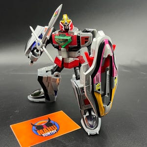 May include: A red, silver, and black robot action figure with a sword and shield. The figure is in a battle-ready pose, with a shield on its left arm and a sword in its right hand. The figure has a green chest plate and a yellow head crest. A small orange card with the text "MAHU SUNTOY" is in the foreground.
