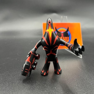 Puede incluir: Figura de acción negra con detalles rojos y un diseño de estrella amarilla en el pecho. La figura tiene un accesorio en forma de arma en un brazo y posa con un brazo extendido. Un logotipo es visible en el fondo.