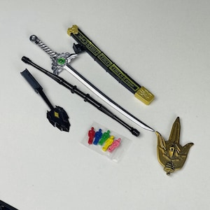 以下が含まれることがあります： 緑色の宝石が付いた銀色の剣、黒い杖、金の盾、小さなカラフルなフィギュアなど、おもちゃの武器とアクセサリーのコレクション。剣には、金のアクセントが付いた黒い鞘が付いています。