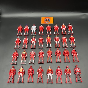 Peut inclure: Une collection de petites figurines rouges avec divers accents colorés. Chaque figurine est debout, les bras le long du corps. Certaines figurines ont des détails blancs, jaunes ou noirs. Les figurines sont disposées sur une surface sombre.