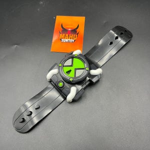 May include: A black and green toy watch with a circular face and a black strap. The watch face has a green and black design with white accents. The watch is a toy from Mahu Suntoy.