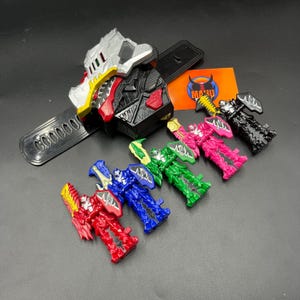 Puede incluir: Un reloj de juguete negro y plateado con una correa negra y una serie de figuras de juguete coloridas. Las figuras son rojas, azules, verdes, rosas y negras. El reloj tiene una serie de botones y un acento rojo.