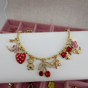 May include: Gold charm bracelet with pendants such as a red strawberry, teddy bear, cherries, and a tree. The bracelet is displayed on a white surface, with a pink jewellery box in the background.
