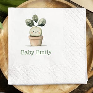 May include: White square paper napkin with a watercolor illustration of a smiling plant in a pot. The text "Baby Emily" is printed in green below the illustration. The napkin has a textured, quilted pattern.