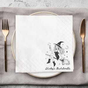May include: A white square paper napkin featuring a black line art illustration of a witch riding a broomstick while holding a champagne flute, surrounded by stars and a crescent moon, with the text Shirley's Bachelorette printed below.