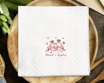 May include: White square paper napkins with a diamond pattern. The napkins feature a pink and gold illustration of two wedding rings, a pink ribbon, and the names "David + Sophia" in pink script. Perfect for a wedding.