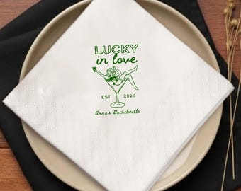 Op de afbeelding: Witte vierkante papieren servet met een groene illustratie van een vrouw in een martiniglas, met de tekst "LUCKY in love" en "Anna's Bachelorette". Het servet ligt op een beige bord en een zwart servet.