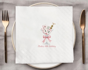 May include: A white square paper napkin featuring a printed illustration of a martini glass filled with mahjong tiles and a small olive skewer. A pink ribbon is tied around the stem of the glass. The text "Madie's 30th Birthday" is printed below.