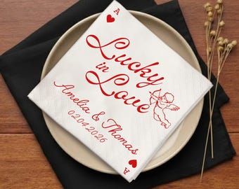 Op de afbeelding: Witte servet met rode tekst "Lucky in Love", namen en een datum. De servet heeft een harten aas en een illustratie van Cupido. De servet ligt op een bord, op een zwarte servet, met gedroogde bloemen.