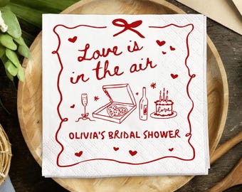 Op de afbeelding: Witte vierkante papieren servet met rode illustraties en tekst. Het servet leest "Love is in the air" en "Olivia's Bridal Shower". De illustraties omvatten pizza, champagne, cake en harten.
