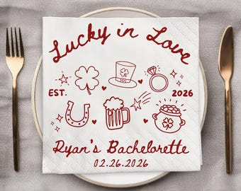 Op de afbeelding: Witte papieren servet met rode tekst en illustraties. De tekst luidt "Lucky in Love" met tekeningen met het thema St. Patrick's Day. De onderkant van het servet luidt "Ryan's Bachelorette" en de datum 02.26.2026.