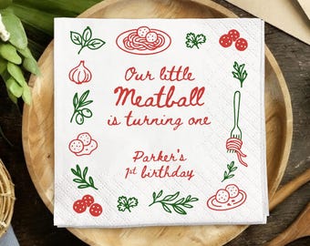 May include: White square paper napkin with red and green illustrations of pasta, meatballs, and herbs. Text reads "Our little Meatball is turning one" and "Parker's 1st birthday". The napkin is on a wooden plate.