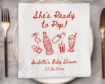 May include: White paper napkin with red text and illustrations. The text reads "She's Ready to Pop!" and "Isabella's Baby Shower" with a date. Illustrations include a pacifier, bottle, soda bottle, drink, and milkshake.