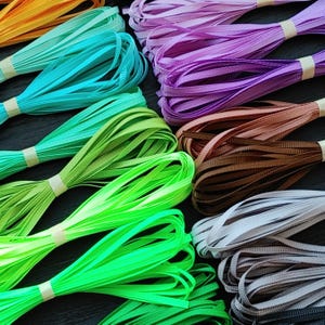 May include: Bundles of colorful satin ribbon in shades of orange, teal, green, purple, brown, and gray. Each bundle is secured with a small piece of tape. The ribbons are arranged on a dark surface.