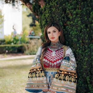 Può includere: Una donna indossa un abito tradizionale ricamato con collo alto e maniche svasate. L'abito presenta accenti rossi, bianchi e blu, con abbellimenti argentati e dettagli di monete. Le maniche sono decorate con motivi oro e neri.