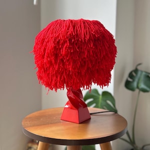Puede incluir: Una vibrante lámpara de mesa roja con una pantalla única y texturizada. La pantalla está hecha de un material denso y peludo, y se asienta sobre una base geométrica roja con un soporte retorcido. La lámpara está colocada sobre una pequeña mesa auxiliar redonda de madera.