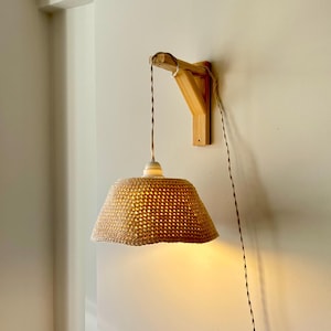 Könnte beinhalten: Ein gewebter beigefarbener Lampenschirm hängt an einer Holzhalterung an einer weißen Wand. Die Lampe ist beleuchtet und strahlt ein warmes Licht aus. Das Stromkabel ist schwarz und weiß. Dekorative Beleuchtung.